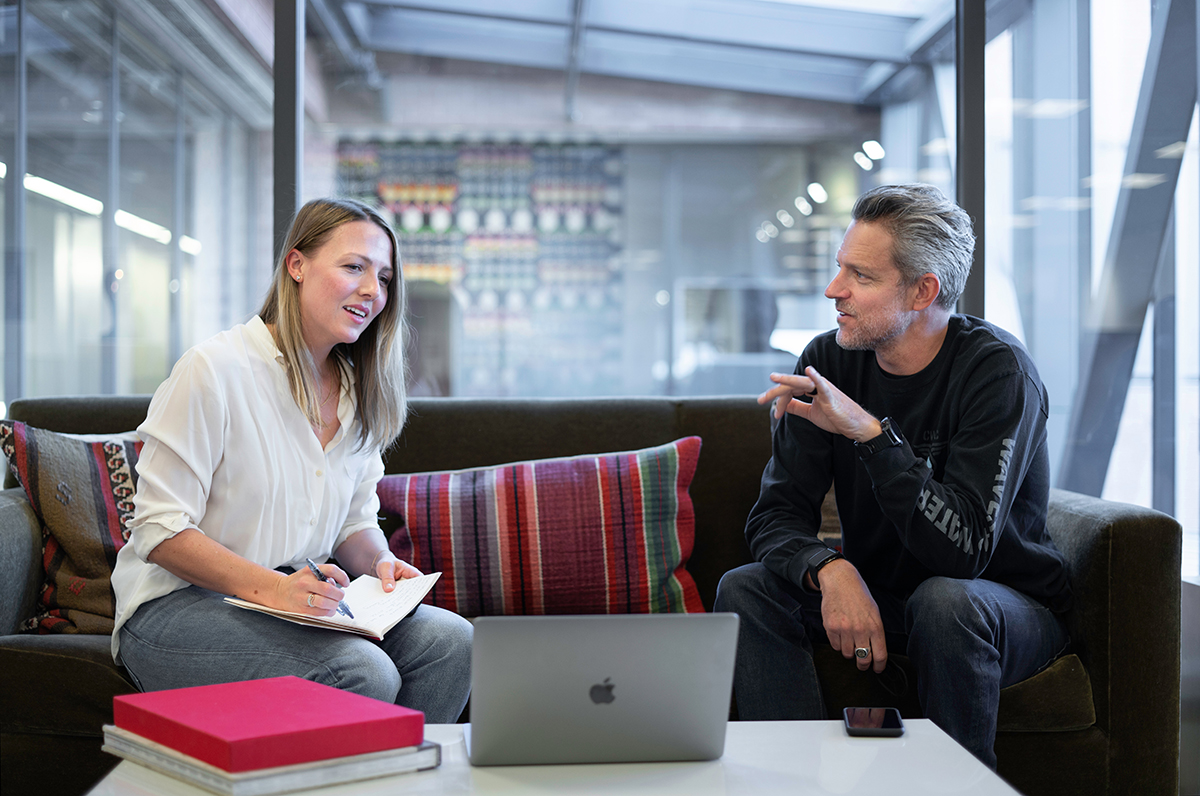 Foto: Unsplash. Kvinna med skrivbok och penna sitter och pratar med en man vid ett bord med en laptop, några böcker och en mobil.